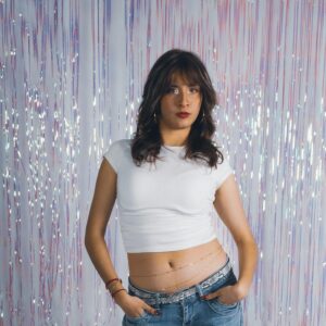 Confident woman in casual attire posing against a shimmering backdrop indoors.